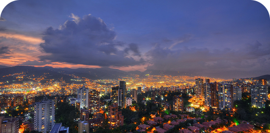 Panorámica nocturna de una ciudad iluminada en un valle, con montañas de fondo bajo un cielo al atardecer.
