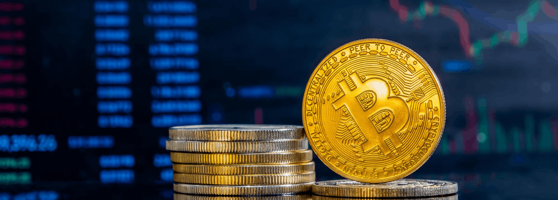 Moneda física dorada de Bitcoin erguida junto a una pila de monedas, sobre un fondo de pantalla con gráficos